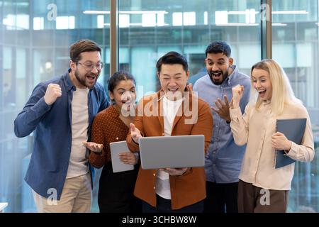 Euphorische Multi-ethnische Geschäftsleute jubeln und geben Gesten, während sie einen Laptop betrachten und feiern ein erfolgreiches Projekt oder eine erfolgreiche Leistung in einem modernen Stockfoto