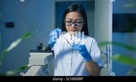 Eine Forscherin in einem Labor trägt Handschuhe und Brille, während sie Proben mit einem Mikroskop unter schwachem Licht analysiert. Stockfoto