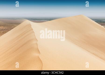 Starker Wind weht Sand über den Kamm einer Düne in der Wüste und schafft wunderschöne Muster auf dem Sand, mit Vegetation und einem Fluss im Hintergrund Stockfoto