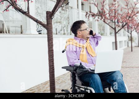 Ein junger Mann im Rollstuhl arbeitet an seinem Laptop, der vor einem wunderschönen Himmel sitzt. Ein behinderter Kerl arbeitet an seinem Laptop am Strand. Funktioniert Remote. Stockfoto