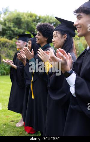 Absolventen in Mützen und Kleidern klatschen fröhlich bei einer Zeremonie im Freien. Abschluss, Feier, akademisch, Erfolg, Leistung, Beginn Stockfoto