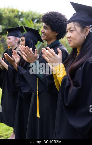 Absolventen in Mützen und Kleidern, die draußen klatschen und Erfolge feiern. Abschluss, Feier, Erfolg, Universität, akademisch, Meilenstein Stockfoto