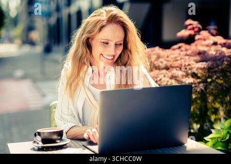 Eine charmante Frau mit blonden Haaren arbeitet draußen an ihrem Laptop und genießt einen sonnigen Tag in einem städtischen Café Stockfoto