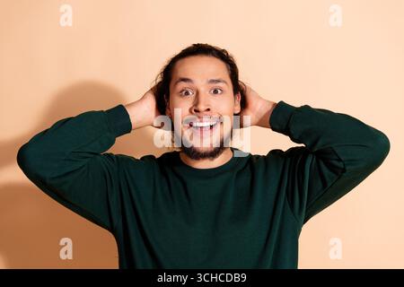 Junger Mann mit fröhlichem Ausdruck vor beigefarbenem Hintergrund, mit grünem Sweatshirt, Hand auf Kopf in Geste Stockfoto