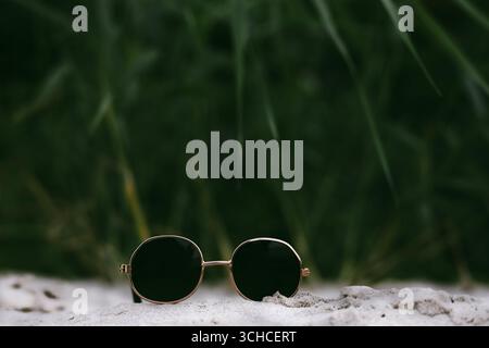 Stilvolle runde Sonnenbrille auf Sand mit verschwommenem grünem Hintergrund. Eine runde Sonnenbrille mit schwarzer Brille liegt auf einer sandigen Oberfläche, mit grüner Farbe Stockfoto