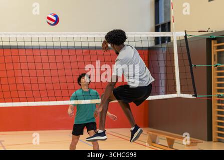 Verschiedene männliche Freunde, die Volleyball über das Netz im Schulstudio mit Bank und Kletterstangen sammeln. Leichtathletik, Teamwork, aktiv, klug, wettbewerbsfähig, leis Stockfoto