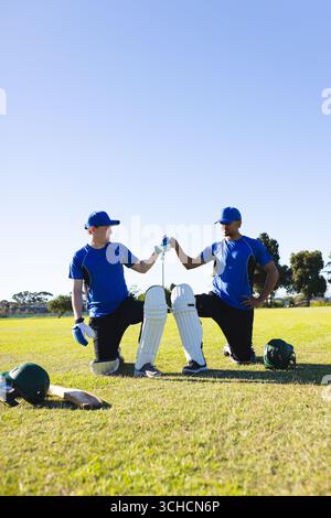 Verschiedene männliche Teamkollegen knieten und Faustschläge auf dem Cricketfeld in blauen Trikots mit Fledermaus und Helmen. Athleten, Teamwork, Sport, Outdoor, Sieg, Cama Stockfoto