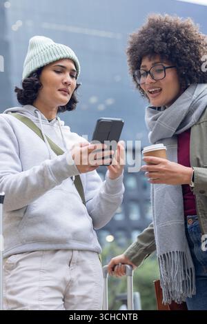 Verschiedene Freundinnen stehen auf dem Bürgersteig, um das Smartphone zu überprüfen und Kaffeetasse und Koffer in der Hand zu halten. Urban, Freundschaft, lässig, lebhaft, Lifestyle, Co Stockfoto