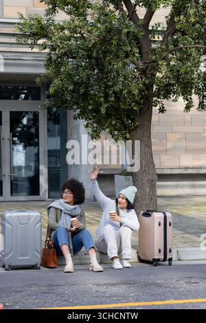 Verschiedene weibliche Freunde sitzen auf der Bordsteinkante vor dem Büro und trinken Kaffee neben Koffern und Handtaschen. Freizeit, Reisen, Freundschaft, Urban, Lifestyle, Modus Stockfoto