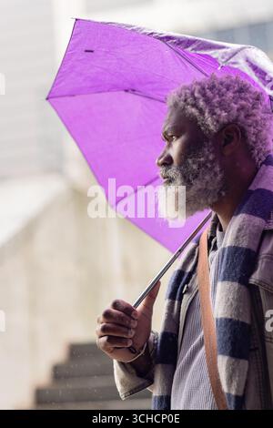 Reifer afroamerikanischer Mann auf dem plaza, der lila Schirm hält, Umhängetasche trägt, Kopierraum. Urban, Architektur, Wetter, Mode, Reife, Fil Stockfoto