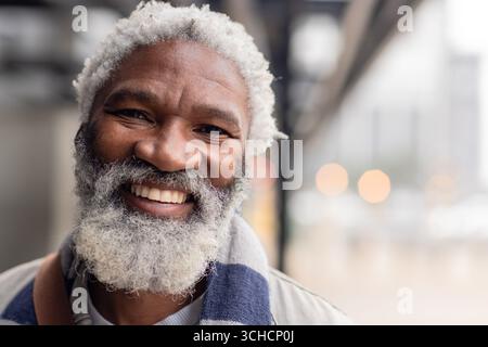 Afroamerikaner lächelt, während er einen gestreiften Schal und eine leichte Jacke auf dem Bürgersteig trägt. Porträt, Senior, offen, Mode, Lifestyle, Vibr Stockfoto