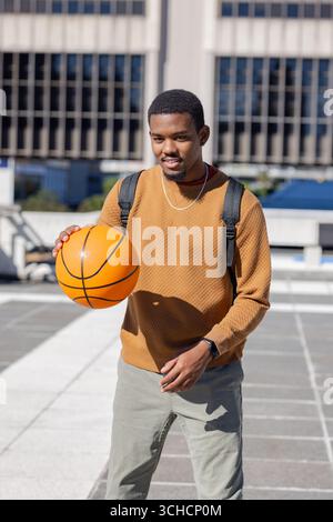 Afroamerikanischer Mann im Sweatshirt, tröpfelt orange Basketball auf dem Dach mit Rucksack, Smartwatch. Urban, sportlich, Freizeit, Sport, Jugend, Stadtbild, Stockfoto