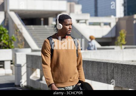 Afroamerikanischer Mann, der auf der plaza läuft, Kopfhörer und Halskette trägt, Skateboard-Rucksack. Urban, Lifestyle, modern, lässig, Outdoor, Stockfoto