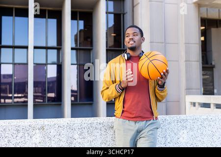 Afroamerikanischer Mann lehnt sich draußen mit Basketball und Kopfhörern an die Wand, Kopierraum. Urban, jugendlich, lässig, lebhaft, energiegeladen, modern, R. Stockfoto