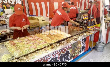 Das Personal an einem geschäftigen japanischen Festivalstand bereitet Okonomiyaki auf einer großen Grillplatte zu. Stockfoto