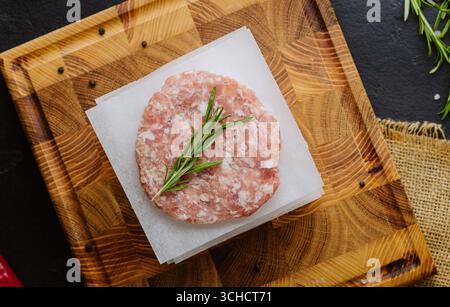 Rohe Burger-Patties mit Pergament und Rosmarin auf einem Holzbrett, fertig zum Kochen Stockfoto