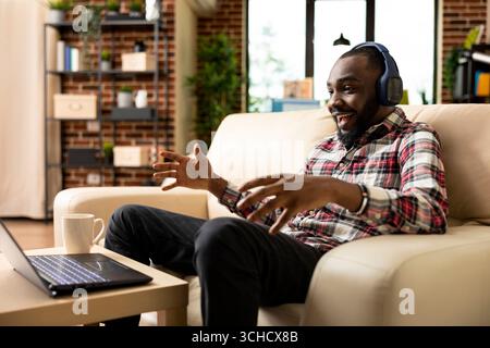 Begeisterter Geschäftsmann mit Kopfhörern zu Hause, der Videochats mit internationalen Arbeitskollegen genießt. Fröhlicher schwarzer Mann, der Filme ansieht, Inhalte streamt oder Musik auf dem Notebook anhört. Stockfoto