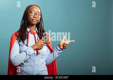 Die afroamerikanische Frau im roten Superhelden-cape zeigt sich mit Zeigefingern zur Seite und steht vor isoliertem Hintergrund mit neutralem Ausdruck. Ernstes schwarzes weibliches Individuum, das zur Seite zeigt. Stockfoto