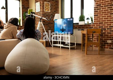 Die jungen Leute sitzen bequem in einem modernen Apartment und streamen beliebte Filme auf einem großen Fernseher. Verschiedene Freunde treffen sich im Wohnzimmer mit Ziegelwänden, um beim actiongeladenen Videospielturnier teilzunehmen. Stockfoto