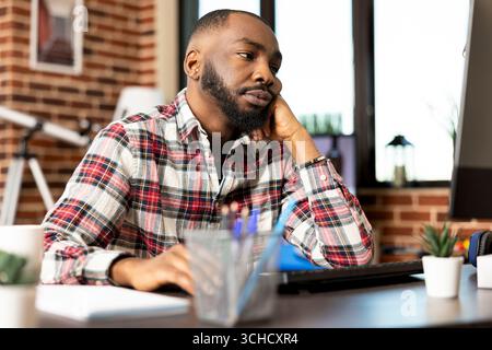 Ein afroamerikanischer Geschäftsmann sitzt am Schreibtisch im Heimbüro, lehnt sich mit müdem Ausdruck auf den Tisch und liest Unternehmensberichte. Bärtiger männlicher Angestellter mit Anzeichen von Langeweile bei Fernarbeiten. Stockfoto