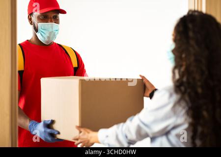 Schwarzer Liefermann in Maske, der der Frau einen Karton gab Stockfoto