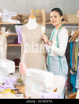 Erfahrene junge Modedesignerin, die mit Stoff und Schaufensterpuppe arbeitet, während sie in der Nähwerkstatt modisches Kleid nähen Stockfoto