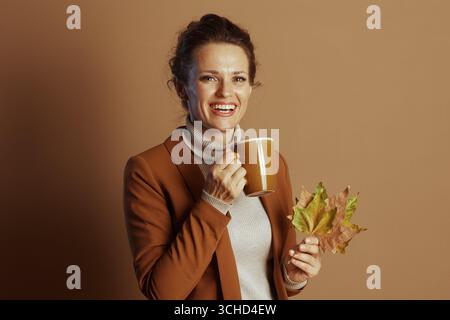 Eine lächelnde Geschäftsfrau in einem braunen Blazer und Rollkragen hält einen gelben Becher und Herbstblätter und vermittelt eine fröhliche, zufriedene und gemütliche Herbststimmung A Stockfoto
