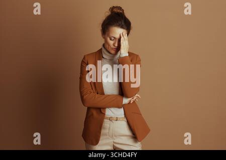 Eine professionelle Frau, gestresst, drückt ihre Hand auf die Stirn und vermittelt Müdigkeit oder Kopfschmerzen. In einem braunen Blazer und beigem Rollkragen gekleidet, s Stockfoto