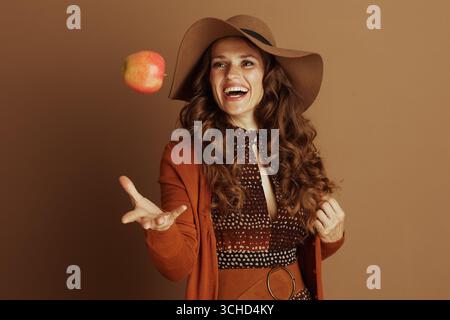 Eine verspielte Frau in breiter Krempe und orangefarbener Strickjacke lächelt und wirft verspielt roten und gelben Apfel in die Luft. Ihre sorglose Pose gegen warme br Stockfoto