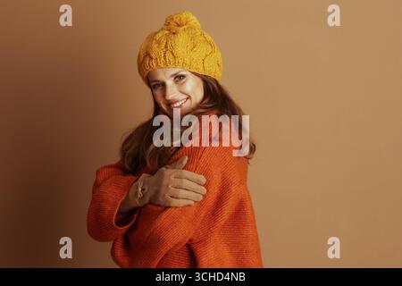 Eine lächelnde Frau mit langen braunen Haaren, trägt eine gelbe Bommelmütze und einen übergroßen orangen Pullover, steht mit überkreuzten Armen und strahlt eine freundliche, Stockfoto