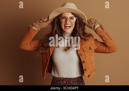 Eine fröhliche Frau mit gewelltem braunem Haar und Brille lächelt, während sie ihren breitkrempigen Hut anpasst, braune Handschuhe, eine orange Jacke und ein gemustertes s trägt Stockfoto
