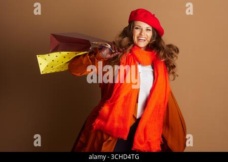 Eine fröhliche Erwachsene Frau in roter Baskenmütze, orangefarbenem Schal und orangefarbenem Trenchcoat lächelt breit in die Kamera. Sie hält zwei gemusterte Einkaufstaschen gegen Lichte Stockfoto