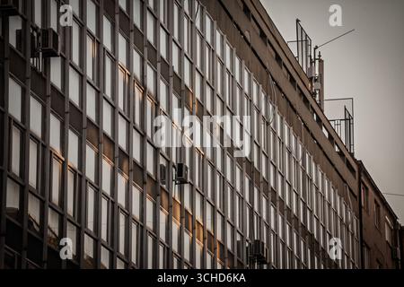 Reihen von reflektierenden Fenstern in einem 1970er-Geschäftsblock in der Dämmerung in Belgrad. Eine modernistische Gitterfassade zeigt sich wiederholende Module, Klimaanlagen und warme Abendlese Stockfoto