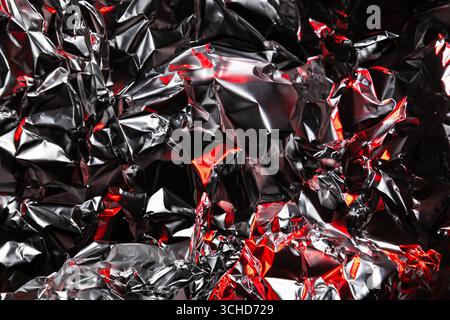 Nahaufnahme einer zerknitterten Aluminiumfolie mit glänzender silberner Oberfläche und dramatischen roten Highlights. Abstrakter metallischer Hintergrund mit reflektierender Textur und Kunst Stockfoto