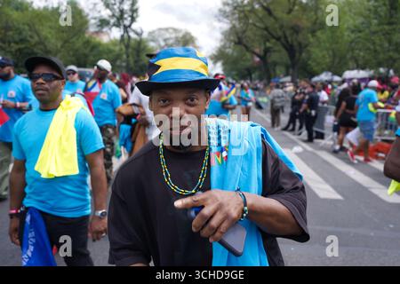 Ein Mann in einem blau-gelb-schwarz gemusterten Hut lächelt in die Kamera, trägt ein schwarzes T-Shirt und ein hellblaues Handtuch über den Schultern. Stockfoto