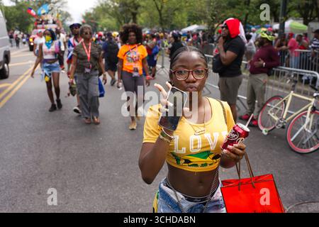 Eine junge Frau in gelbem Oberteil posiert für ein Foto mit einem Handy und einer Dose Dr. Pepper auf einem Straßenfest oder einer Parade. Stockfoto