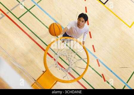Springender asiatischer männlicher Basketballspieler, der den Ball durch das Basketballnetz auf dem Boden mit farbigen Linien platziert Stockfoto