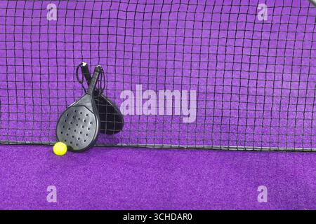Zwei schwarze Padel-Schläger, die sich gegen ein schwarzes Netznetz lehnen, auf einem violetten synthetischen Platz mit gelbem Ball Stockfoto