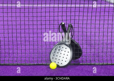 Zwei Padel-Schläger, die sich gegen ein schwarzes Netznetz auf dem purpurfarbenen Rasenplatz mit gelbem Ballkontrast lehnen Stockfoto