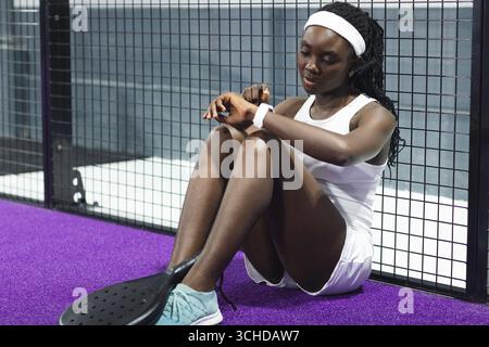 afroamerikanerin in weißem Outfit, die Smartwatch auf einem violetten Padel-Court mit Schläger überprüft Stockfoto