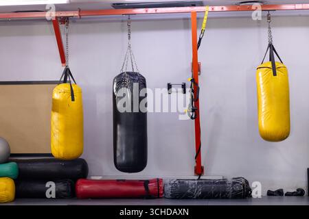 Drei Boxsäcke hängen vom roten Balken an der grauen Wand des Fitnessstudios neben Matten, trx-Riemen Stockfoto