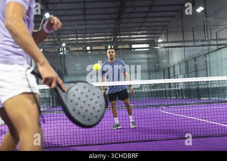 Zwei Männer spielen Padel-Tennis auf einem violetten Platz in der Sporthalle mit Schlägern und gelbem Ball Stockfoto