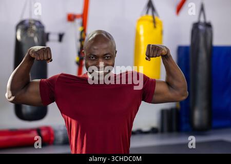 afroamerikanischer Mann, der Bizeps biegt, lächelt im Boxstudio mit schwarzen und gelben Boxsäcken Stockfoto