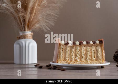 Ein wunderschön präsentiertes Stück Kuchen auf einem weißen Teller, begleitet von einer weißen gerippten Vase mit getrocknetem Pampasgras, auf einem neutralen Rücken Stockfoto