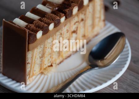 Nahaufnahme einer Kuchenscheibe mit Schokoladen- und Sahneschicht auf einem weißen Teller mit einem Löffel, die die reichhaltigen Texturen des Kuchens und das köstliche Zuckerguss hervorhebt Stockfoto