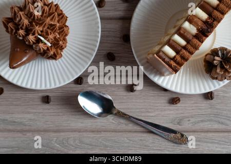 Nahaufnahme von zwei Desserts, einer igelförmigen Schokoladenspeise und einem mehrschichtigen Kuchen, auf weißen Tellern mit verstreuten Kaffeebohnen und einem Löffel auf einem rustikalen wo Stockfoto
