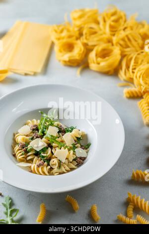 Fusilli-Pasta-Gericht mit Parmesan, Rucola und Hackfleisch, serviert auf einem weißen Teller, umgeben von anderen Nudelsorten Stockfoto
