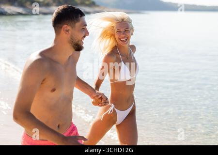Ein Paar genießt einen verspielten Moment am Strand, hält Hände und lacht, während sie durch flache Wellen von kristallklarem Wasser laufen. Stockfoto