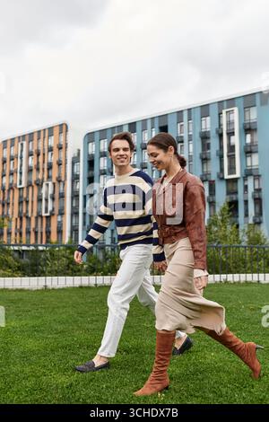 Ein junges Paar schlendert Hand in Hand durch einen grünen Park mit moderner Architektur. Stockfoto