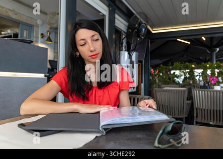 Nachdenkliche Frau liest Menü im Restaurant im Freien. Junge Frau in rotem Oberteil liest nachdenklich ein Restaurantmenü, während sie draußen sitzt und entscheidet, was Stockfoto
